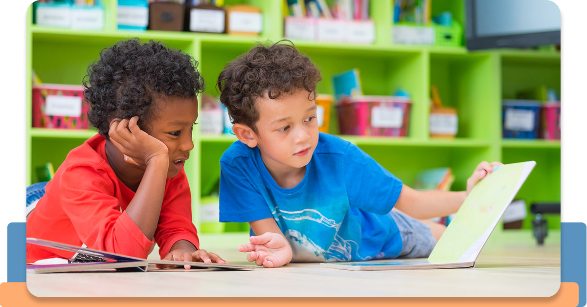 Two children reading books on the floor of a classroom.