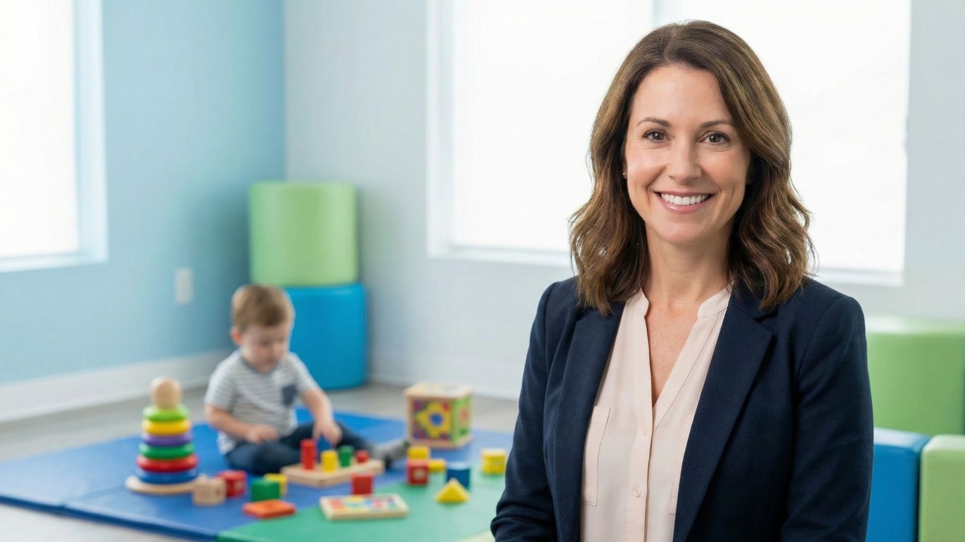 A professional BCBA smiling warmly in a bright, modern pediatric therapy clinic with a child playing in the background.